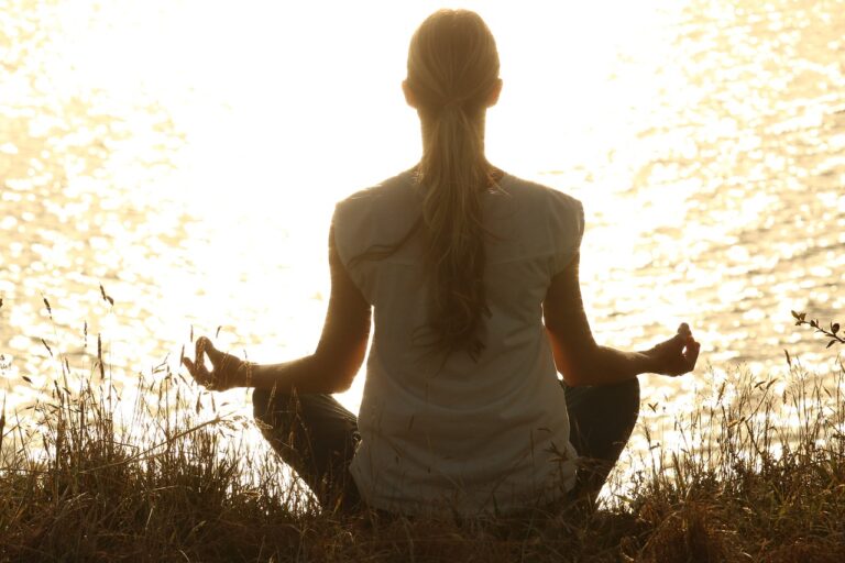 A woman practicing yoga at sunset, seen from behind, in a relaxed pose promoting mindfulness and relaxation.